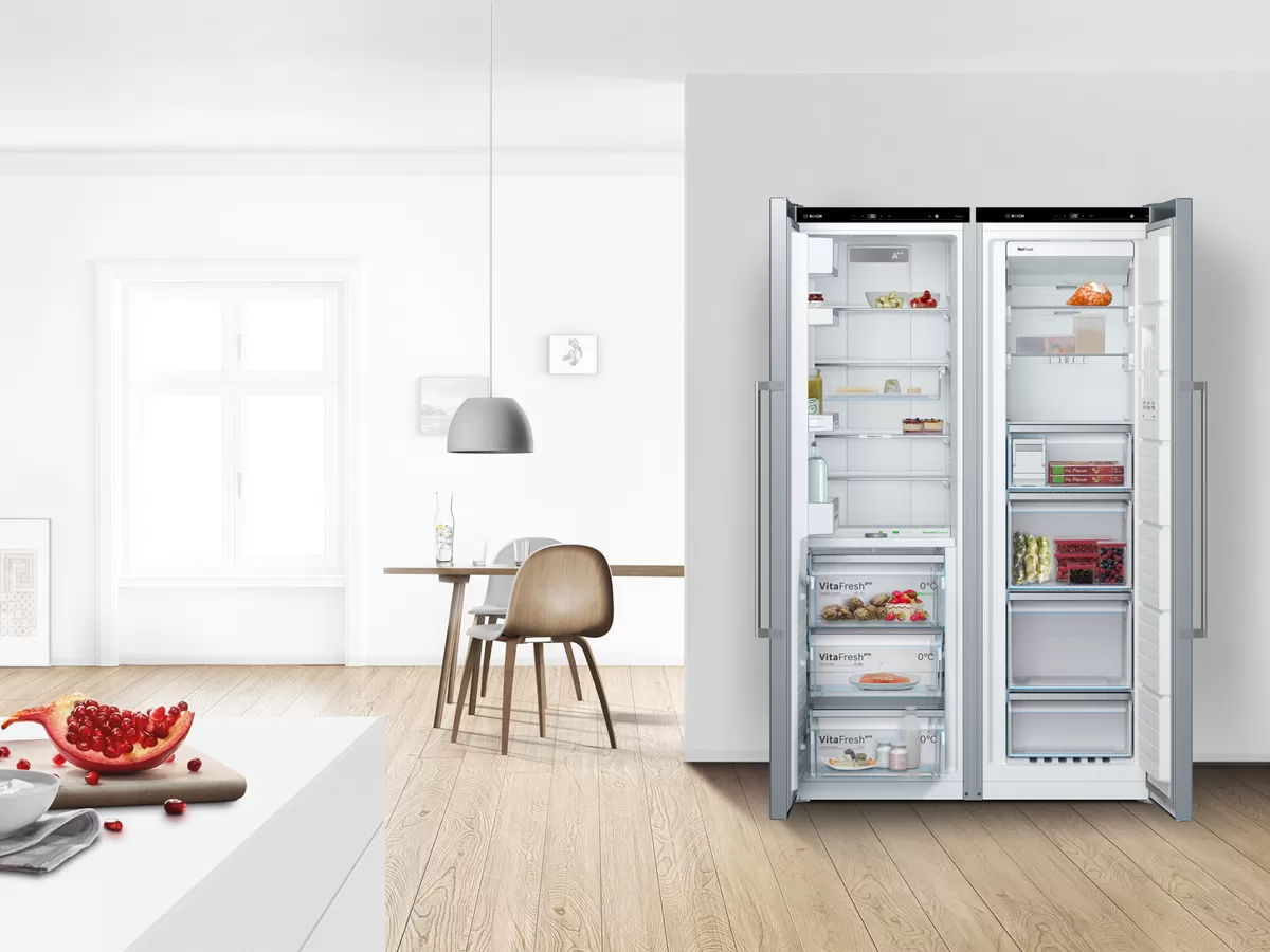 Una moderna nevera abierta llena de alimentos frescos, situada en una cocina luminosa y minimalista con suelos de madera y una sencilla zona de comedor.