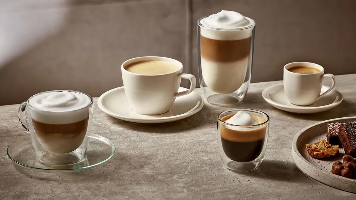 An assortment of coffee drinks in elegant cups, featuring layered lattes, espresso, and a plate of sweet treats on a textured surface.