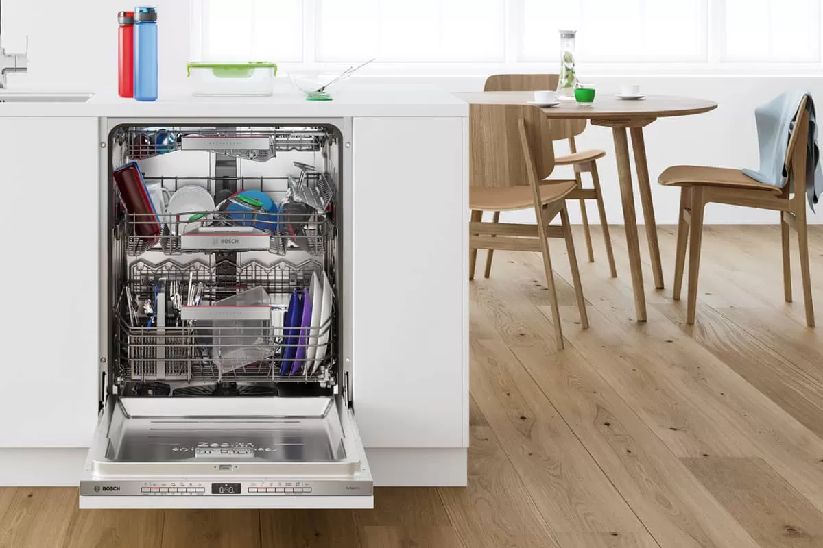 Un lavavajillas Bosch está abierto, lleno de platos limpios, en una cocina moderna con una mesa de comedor de madera y sillas al fondo.