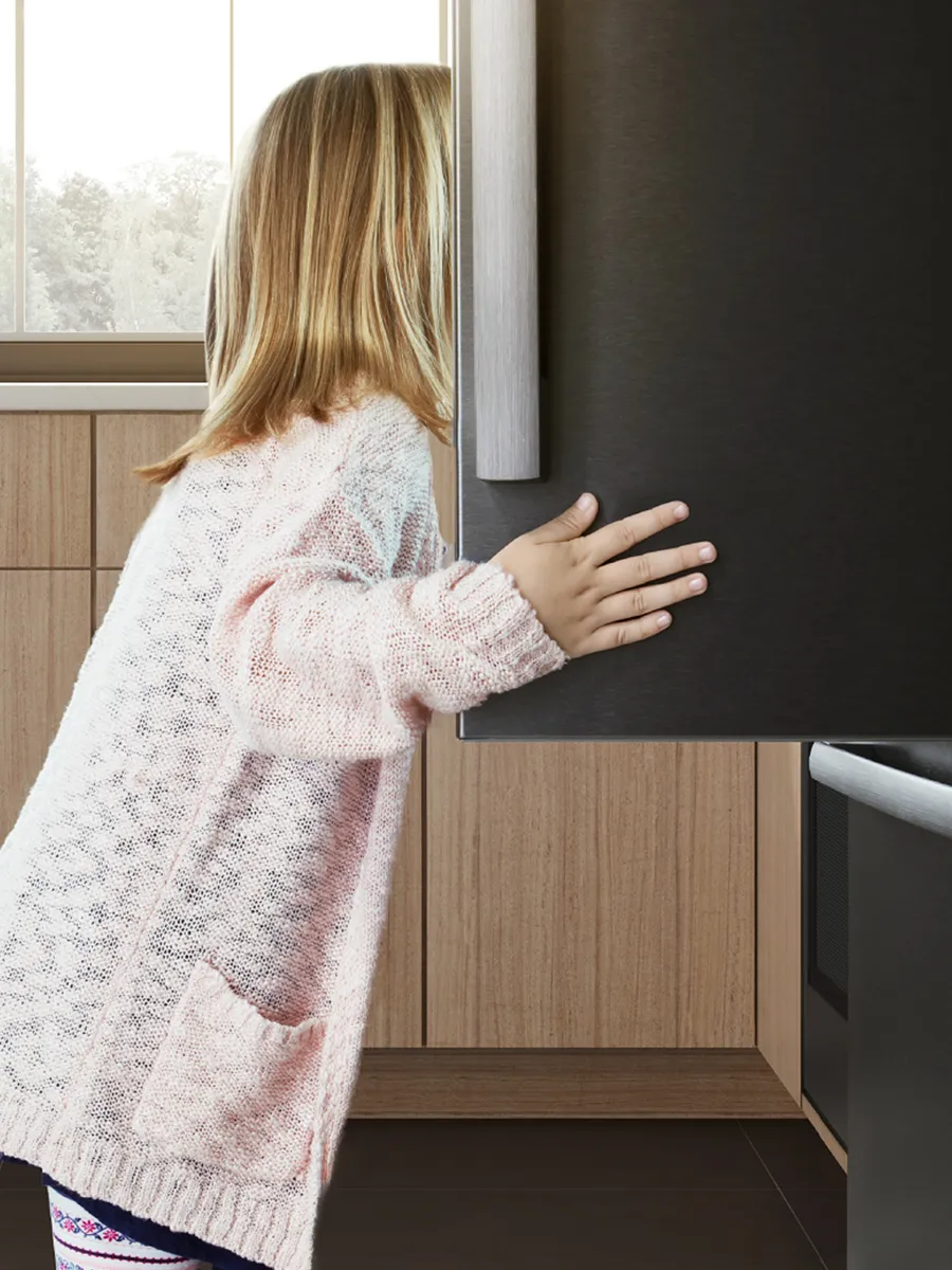 Ein junges Mädchen mit langen Haaren schaut in einen Kühlschrank, ihre Hand ruht auf der Seite, in einer warmen, modernen Küchenumgebung.