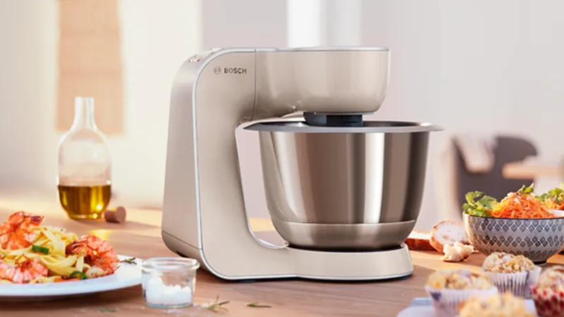 A Bosch kitchen mixer on a wooden countertop with a stainless steel bowl, surrounded by various dishes including pasta, shrimp, and salads, with a bottle of olive oil in the background.