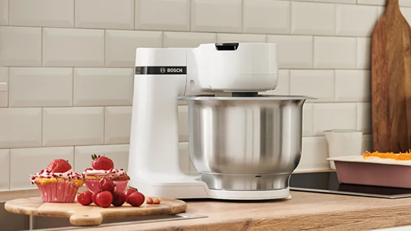 A Bosch kitchen mixer in white with a stainless steel bowl on a wooden countertop, accompanied by pink cupcakes topped with strawberries and a variety of fresh red fruits, with a tiled wall and baking dishes in the background.