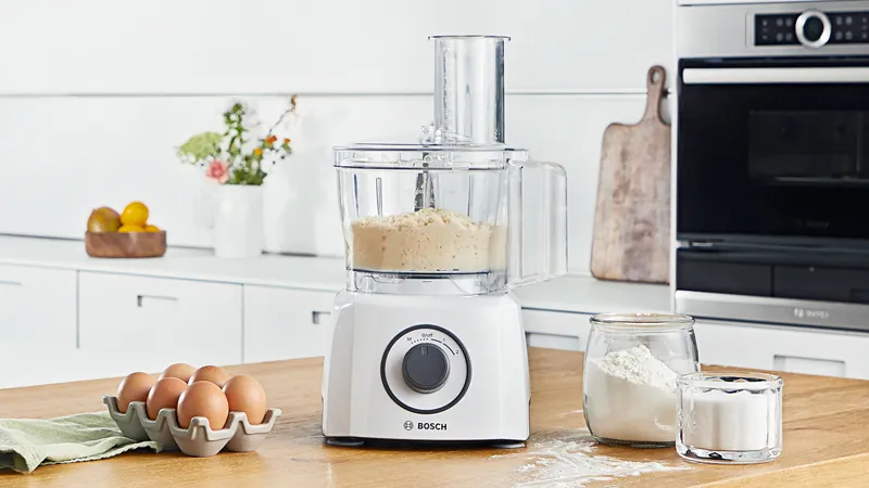 Bosch processor on wood counter with eggs, flour, sugar, and fresh flowers in a vase nearby.