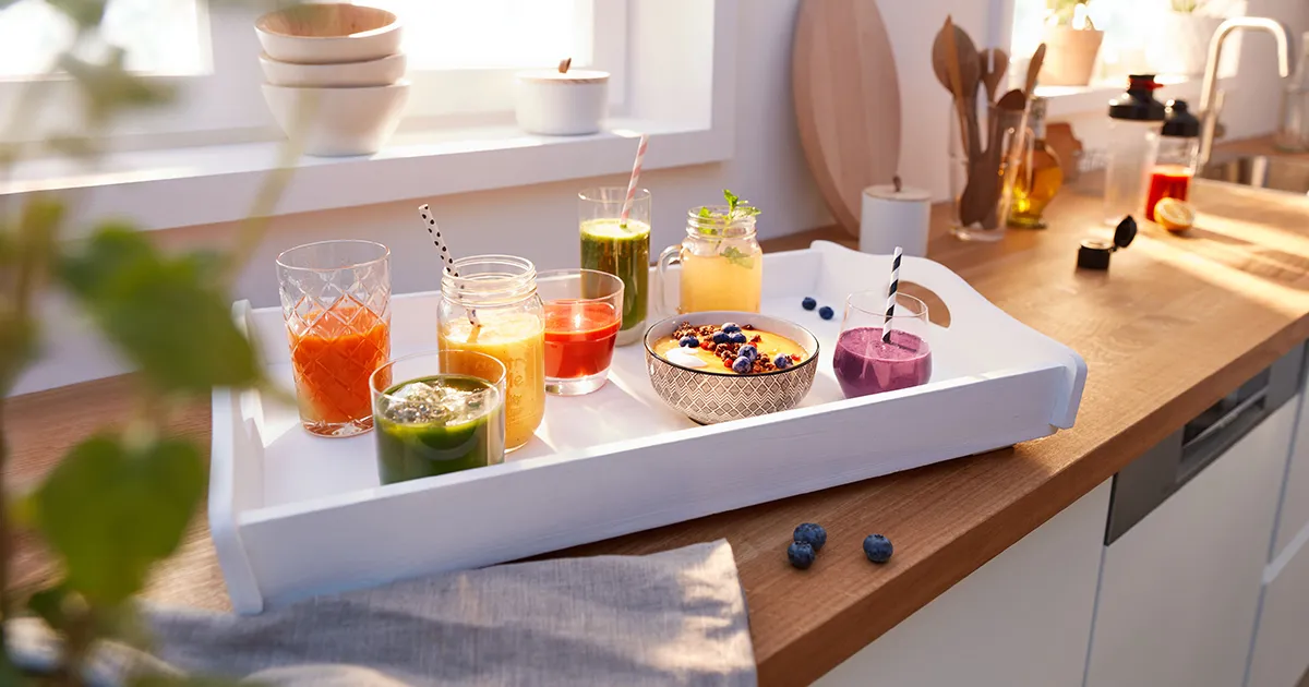 Una elegante bandeja de madera muestra jugos coloridos y un tazón de batido, sobre un fondo de cocina brillante con ingredientes frescos.