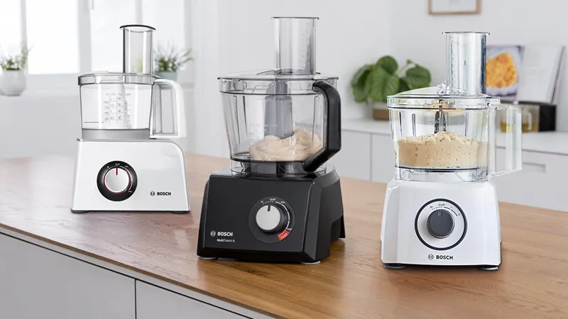 Three Bosch food processors lined up on a wooden countertop: a white food processor on the left, a black food processor in the center with dough inside, and a white food processor on the right containing a preparation, all set against a bright kitchen background with plants.