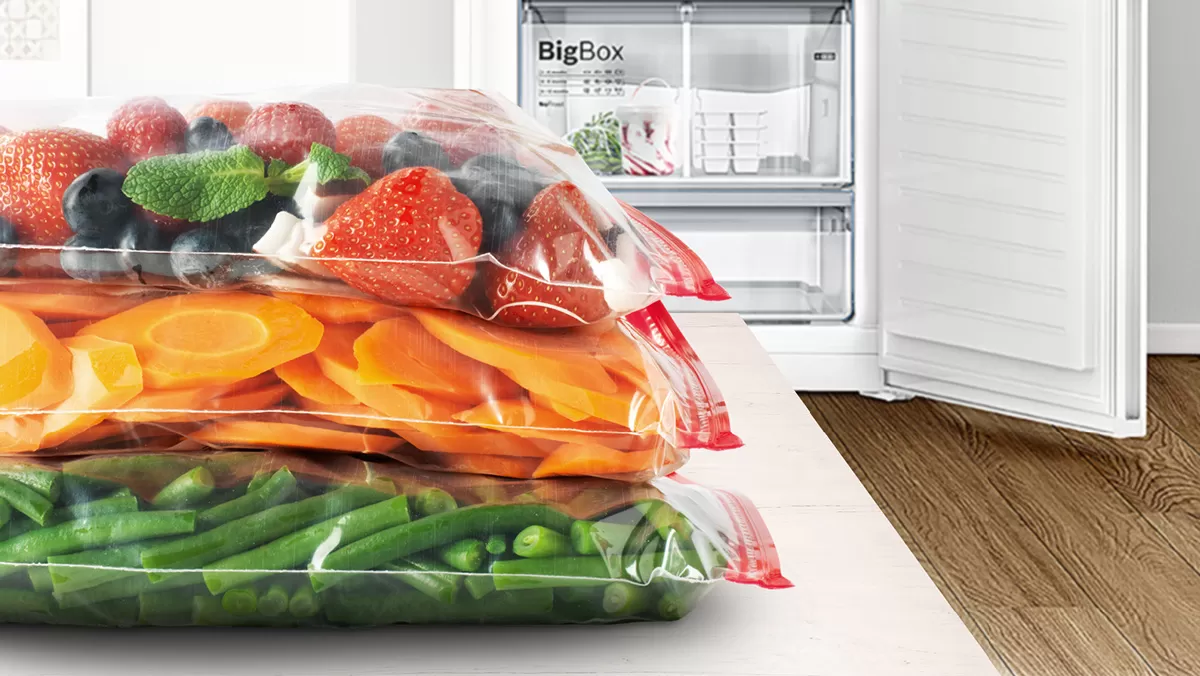 Assorted fruits and vegetables in transparent bags on a table, with an open refrigerator in the background.