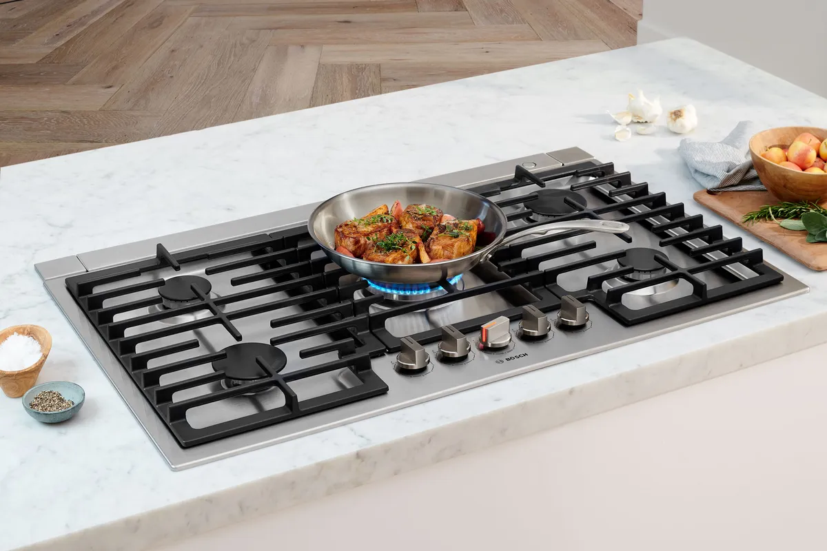 Gas cooktop with burners, pan, and various cooking ingredients on a marble countertop.