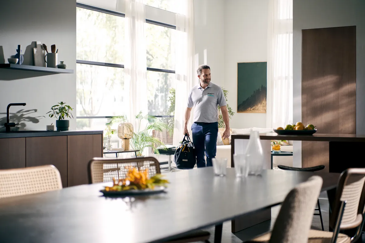 Asmuo įžengia į modernią virtuvę, nešdamas maišelį, o saulės spinduliai patenka per didelius langus, apšviesdami stilingą dekorą.