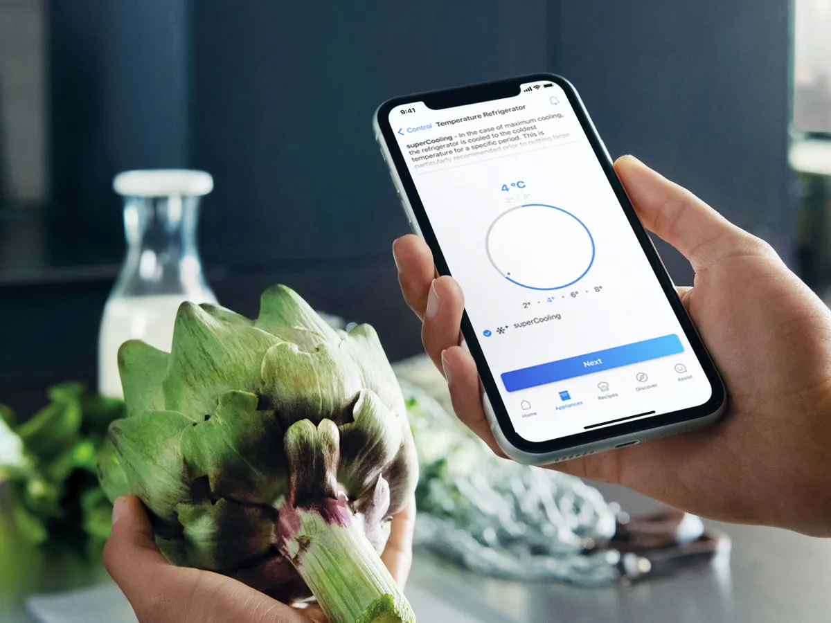 A person holds an artichoke in one hand and a smartphone displaying refrigerator temperature settings in the other, surrounded by fresh produce.