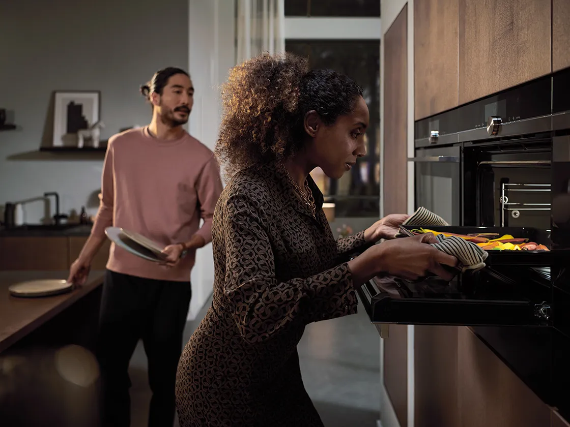 Two people in kitchen, one taking tray out of oven