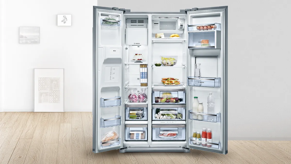 A stainless steel refrigerator stands in a modern kitchen beside a countertop with green limes and a bowl of fruit.