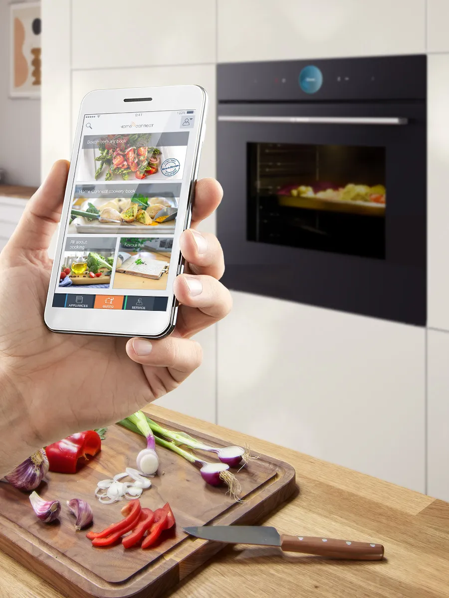 Hand holding smartphone with cooking app; veggies and modern oven on wood counter.