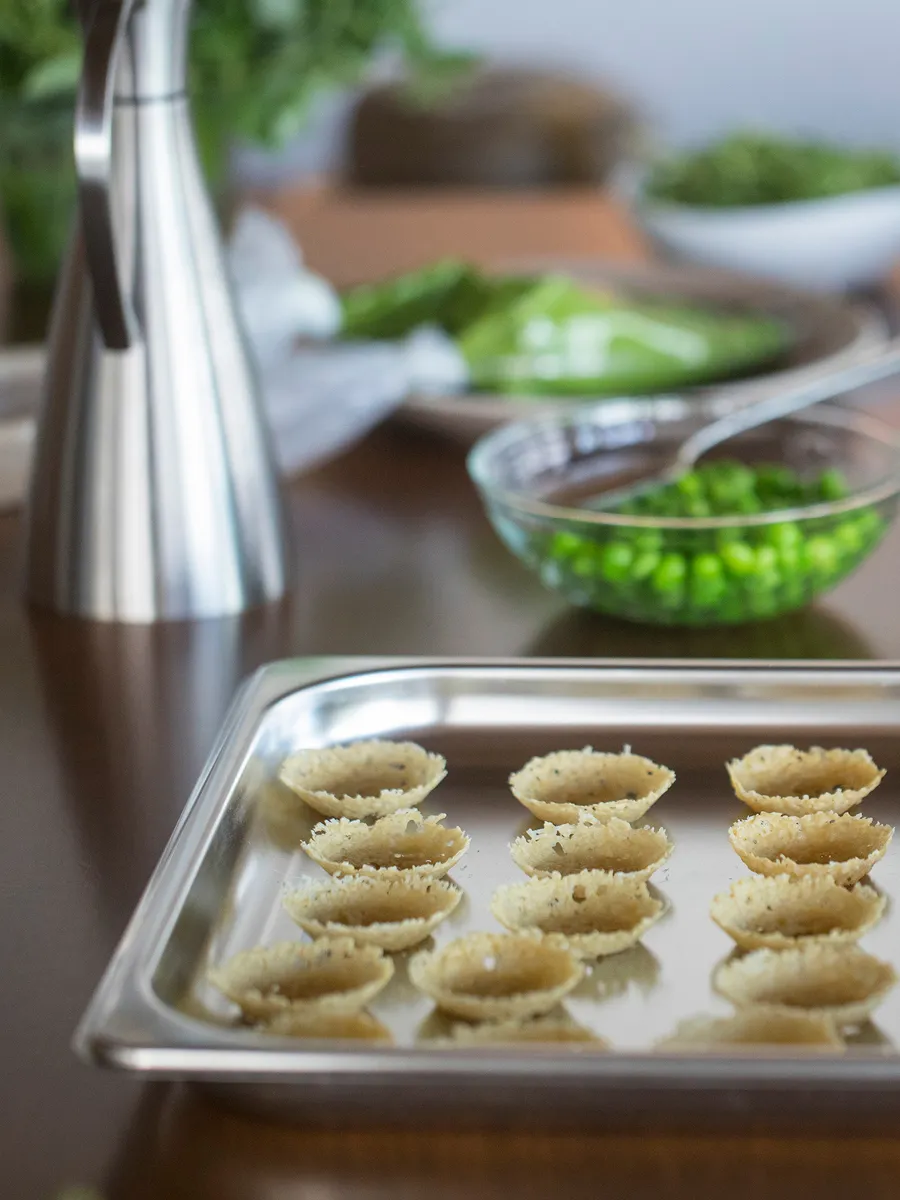 A stainless steel tray holds small, crispy shells, with a glass bowl of green peas and a dressing dispenser in the background.