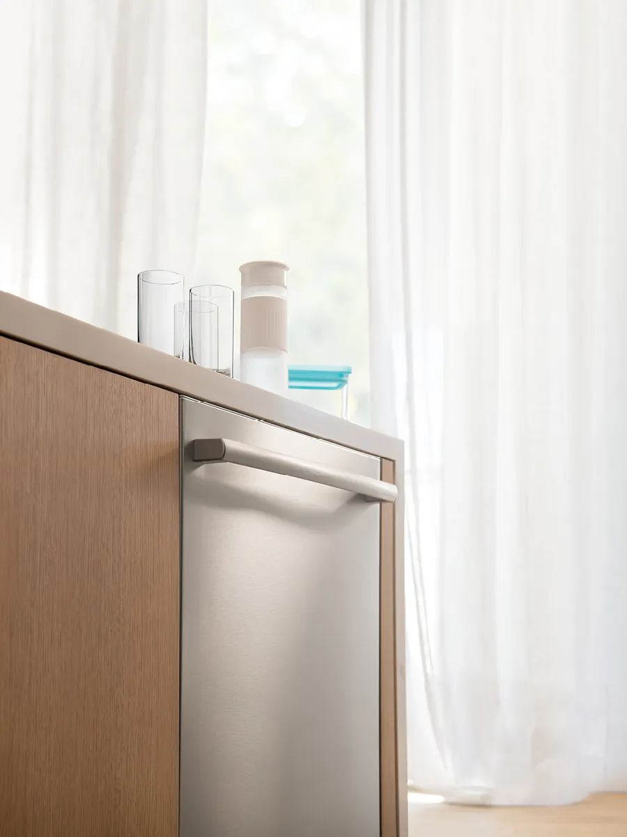 Modern kitchen scene featuring a sleek stainless steel dishwasher, wooden cabinetry, and glasses on the countertop by a bright window.