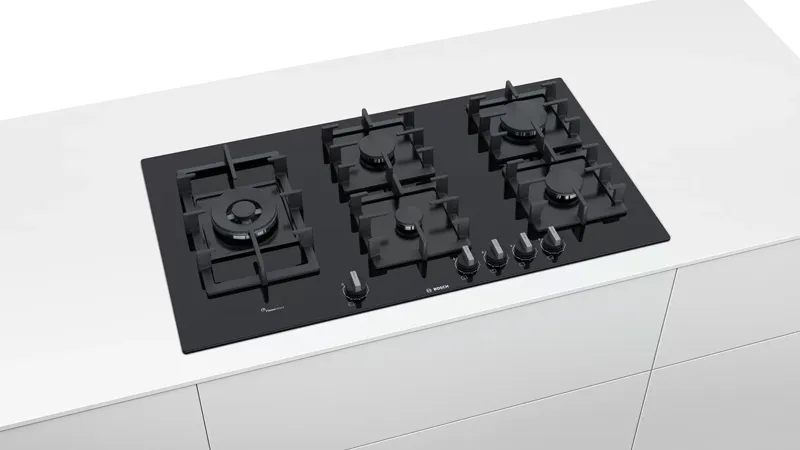 Modern black gas hob with five burners on a sleek white countertop. The design is minimalist, with silver control knobs on the right side.
