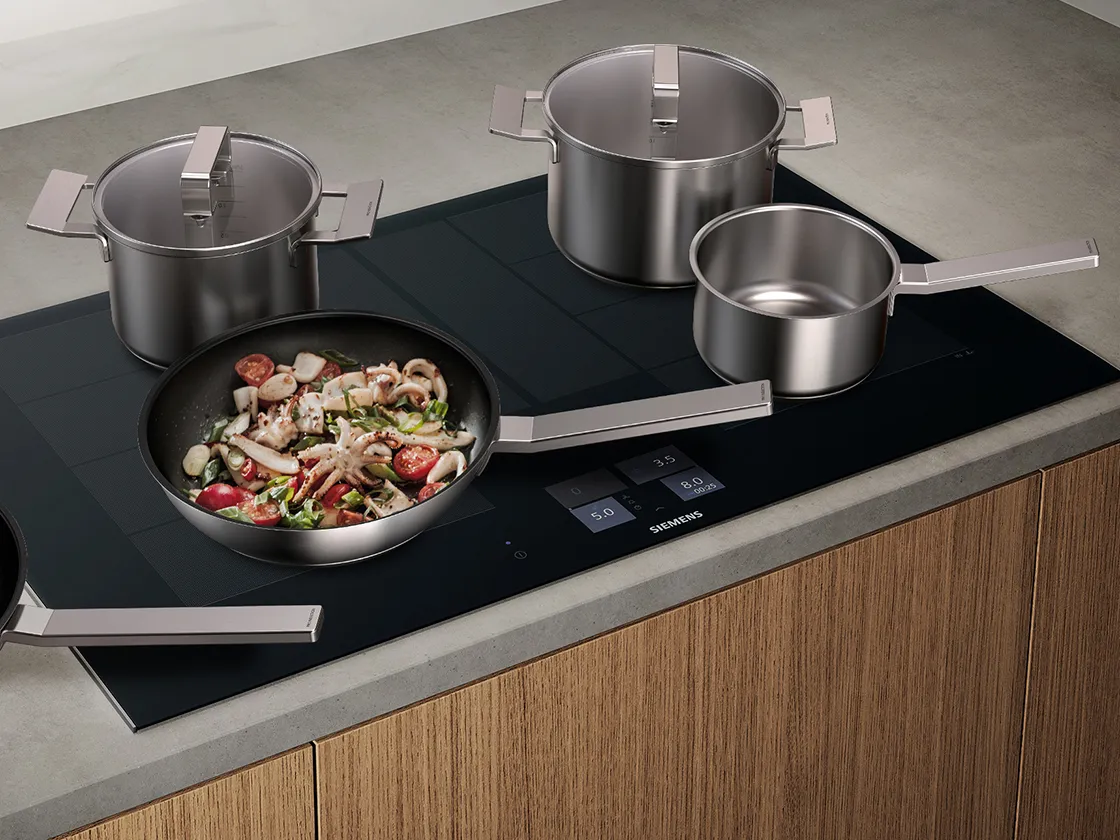 Stainless steel pots and pans on a modern induction cooktop with a vegetable-filled skillet.