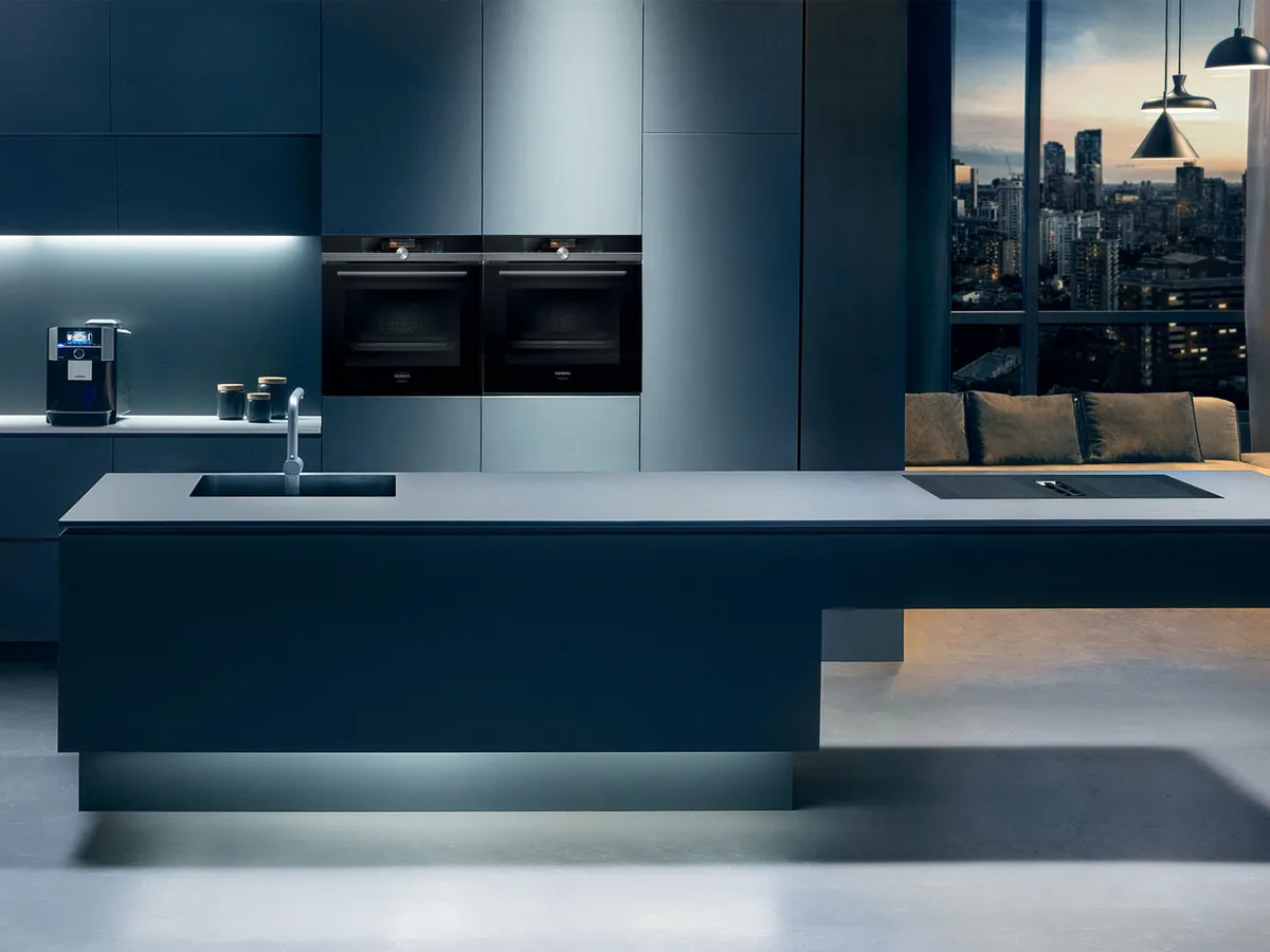 Cuisine moderne avec des meubles sombres et élégants, des plans de travail illuminés, des fours encastrés et une vue sur la ligne d’horizon de la ville à travers de grandes fenêtres.