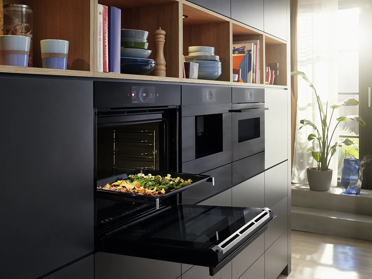 Bucătărie modernă cu dulapuri negre, rafturi deschise, cuptor cu tava de copt, și plante ghiveci lângă fereastră.