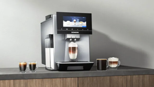 Siemens kavni aparat z ravnokar pripravljenim kavnim napitkom z mlekom in več skodelicami kave okoli na mizi.