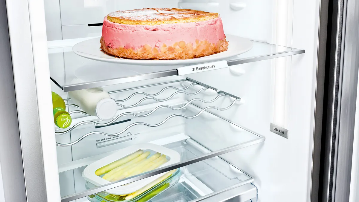 Atidarytas šaldytuvas su pyragu ant lanksčios stiklinės lentynos.