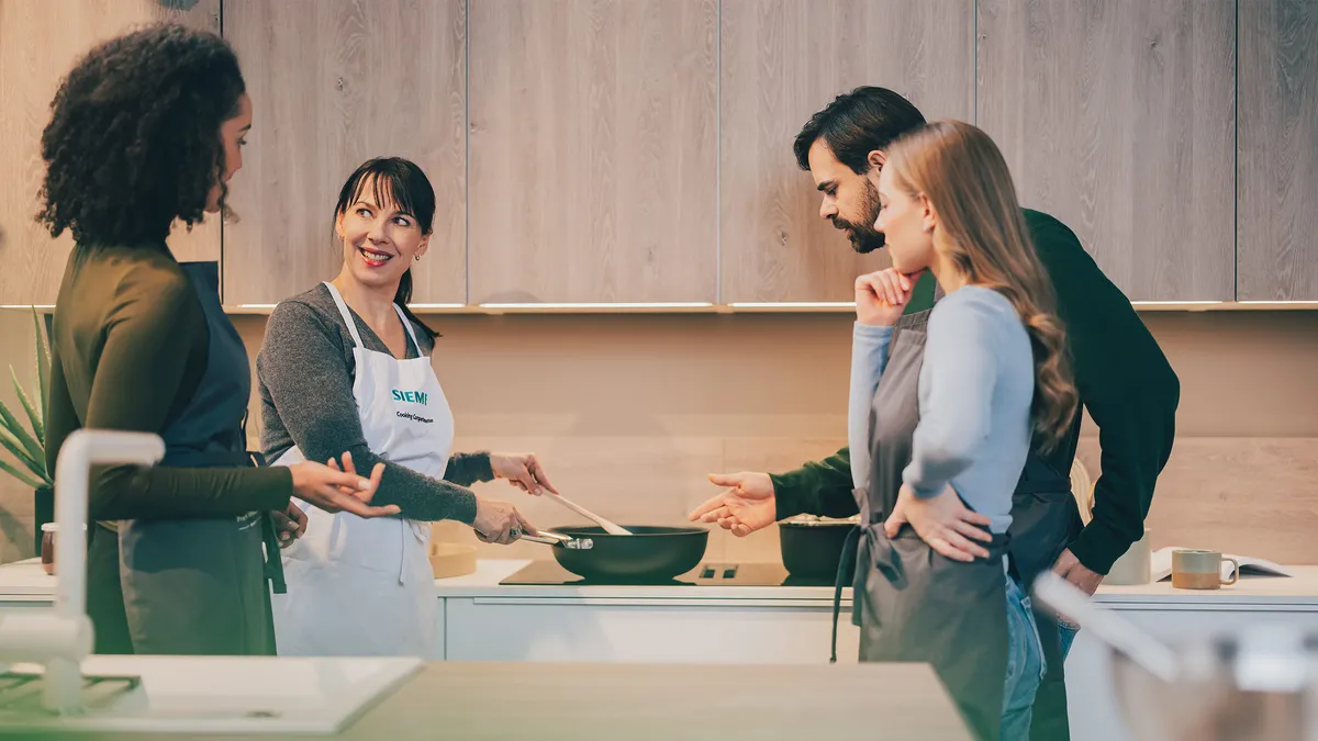 Eine Siemens Expertin erklärt drei Personen die neuesten Funktionen und Anwendungen eines Siemens Kochfelds, während sie mit einer Wokpfanne kocht.