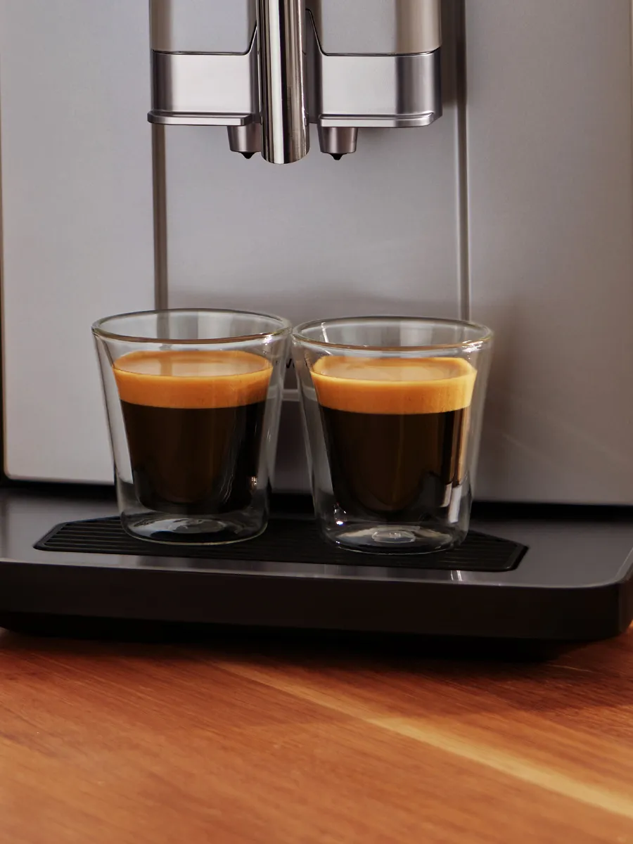 Two glass cups of freshly brewed espresso sit on a sleek coffee machine, showcasing rich crema on top, against a wooden surface.
