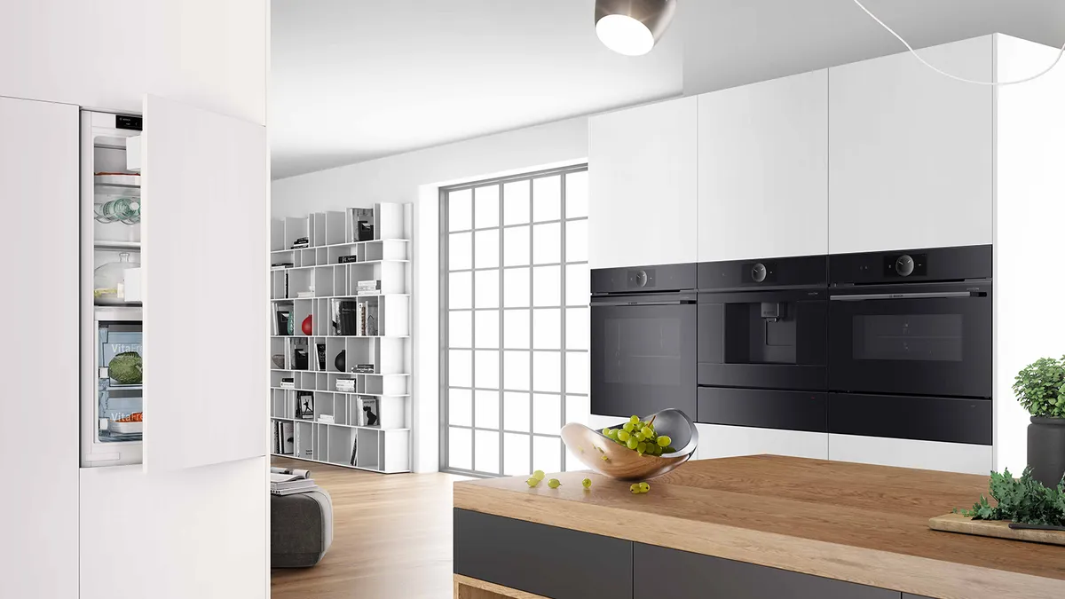 Moderne Küche mit einem eleganten weißen Kühlschrank, eingebauten schwarzen Öfen und einer Holzinsel mit Obstschale und minimalistischem Dekor.