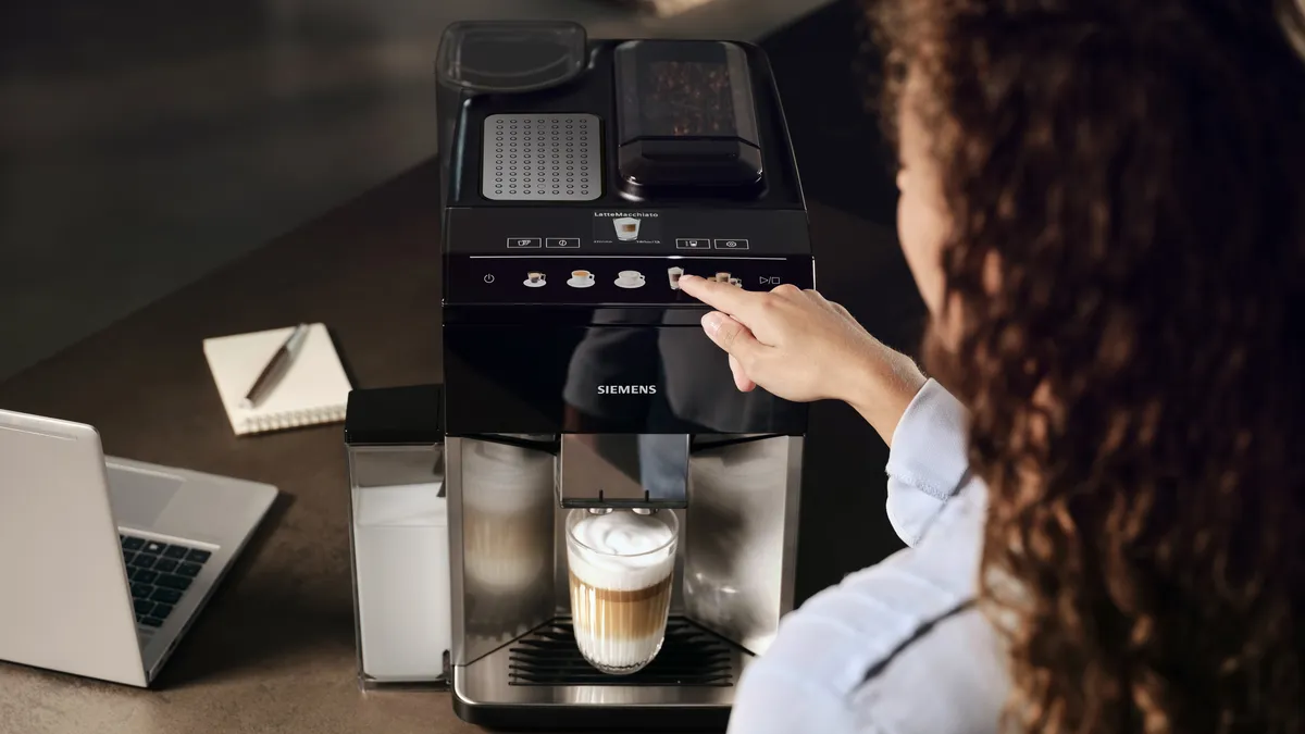 Woman is pressing the cappuccino button at the EQ500 coffee machine, a glass with coffee and milk foam already under the outlet.MASTER | Global | Coffee Machines - 