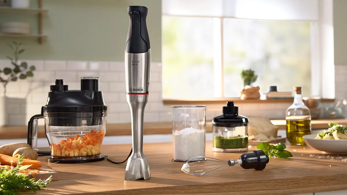 Um liquidificador de mão Bosch e vários acessórios espalhados sobre uma bancada de madeira, juntamente com um copo de leite e outros ingredientes frescos.