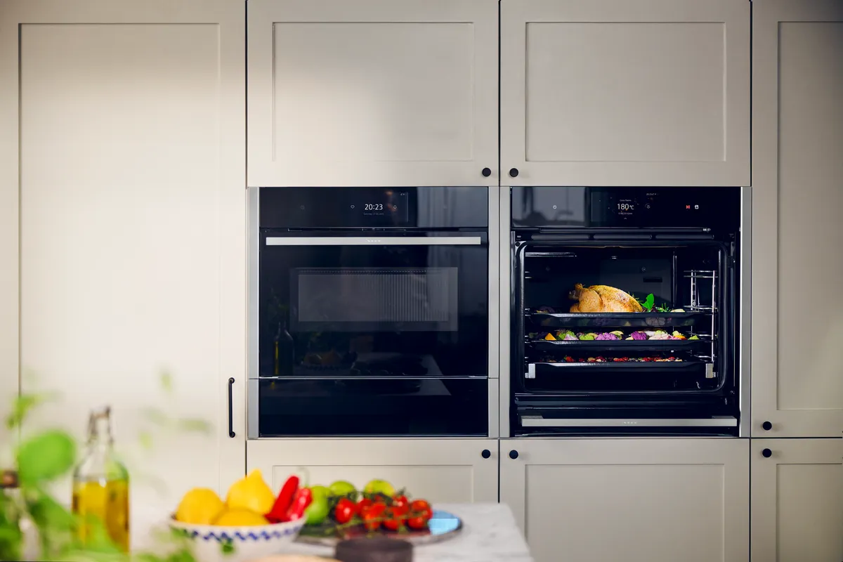 Køkkenet har to indbyggede ovne; den nederste er fyldt med en steg og grøntsager. På bordet er der frisk frugt og olivenolie, hvilket skaber en hjemlig stemning.