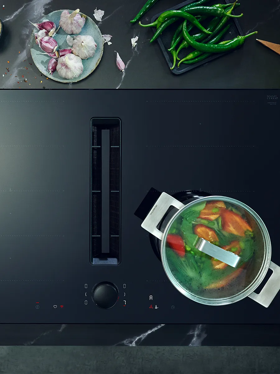 Schwarzes Glaskeramik‑Kochfeld mit mittigem Muldenabzug, rechts ein Topf mit Glasdeckel und kochendem Gemüse (Tomaten, grüne Bohnen/Erbsen), oben links Knoblauch und grüne Chilischoten auf der Arbeitsplatte; Bedienknopf und Anzeige‑Symbole sichtbar.