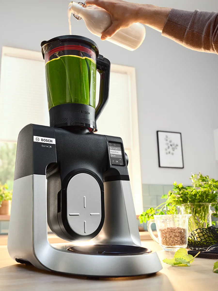 Een persoon giet vloeistof in een Bosch-blender die gevuld is met groene ingrediënten, met een heldere keuken als achtergrond.