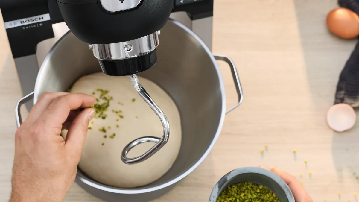 A hand sprinkles chopped pistachios into a mixing bowl of dough, with a Bosch mixer in the background and other baking ingredients nearby.