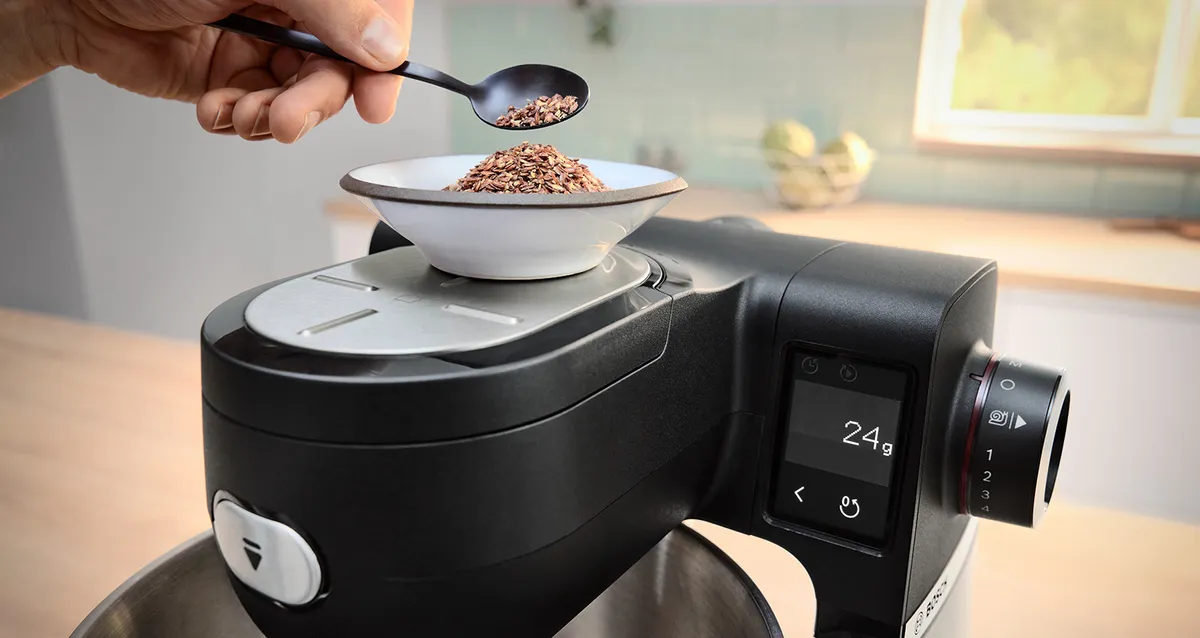 Moedor de café com mão adicionando grãos de café a uma tigela em um balcão de cozinha perto de uma janela. 