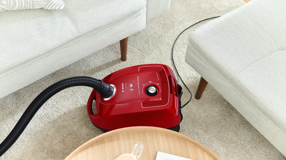 A body of a red, compact cylinder vacuum moves easily through the narrow space separating two couches to clean the carpet.