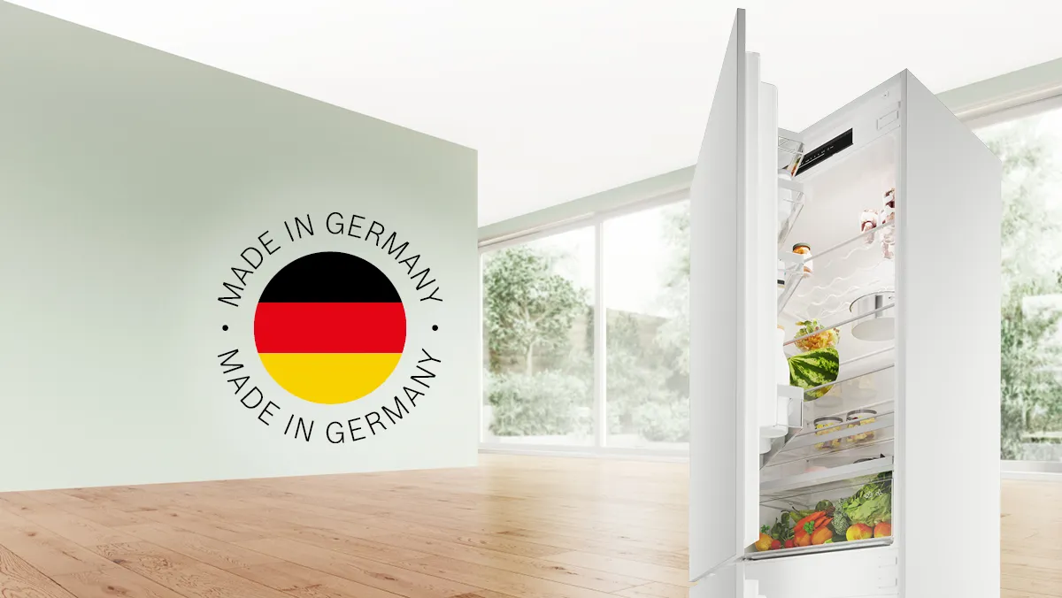 In einem hellen Raum steht ein moderner weißer Kühlschrank offen neben einer Wand mit dem Schriftzug „Made in Germany“ und der deutschen Flagge.