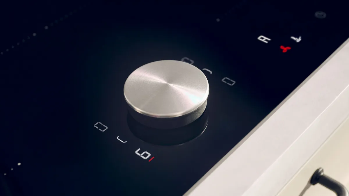 Nahaufnahme eines eleganten, metallischen Bedienknopfes auf einem modernen berührungsempfindlichen Kochfeld, der von digitalen Anzeigen umgeben ist.