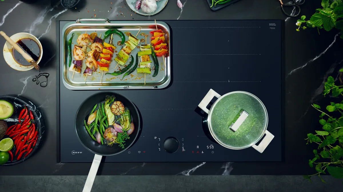 Ein moderner Herd mit gegrillten Spießen, gebratenem Gemüse und einem Topf mit grüner Suppe, umgeben von frischen Zutaten und Kräutern.