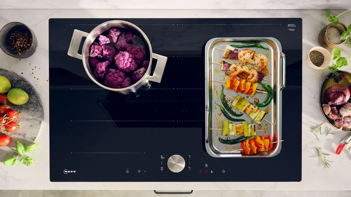 Auf einem modernen Kochfeld stehen ein Topf mit lila Blumenkohl und ein Tablett mit buntem Grillgemüse, umgeben von frischen Zutaten und Gewürzen.