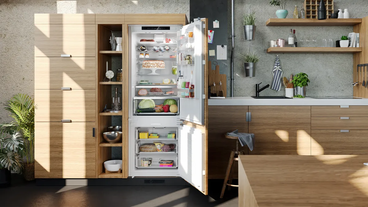 An open built-in fridge freezer inside a light-wood kitchen illuminated by daylight.