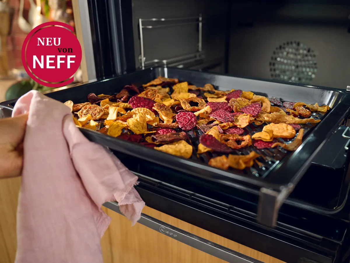 Eine Hand zieht ein Tablett mit bunten, getrockneten Obst- und Gemüsechips aus einem NEFF-Ofen und präsentiert damit gesunde Snackoptionen.