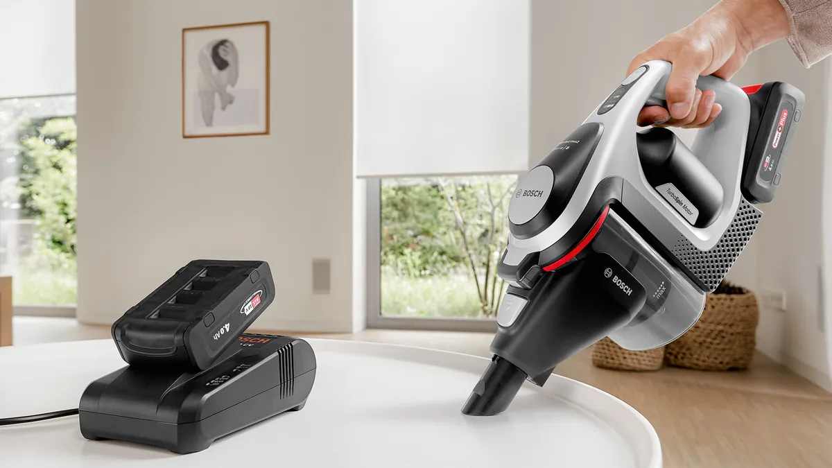 A hand uses an Unlimited vacuum as a handheld to clean a table where a battery charger stands.