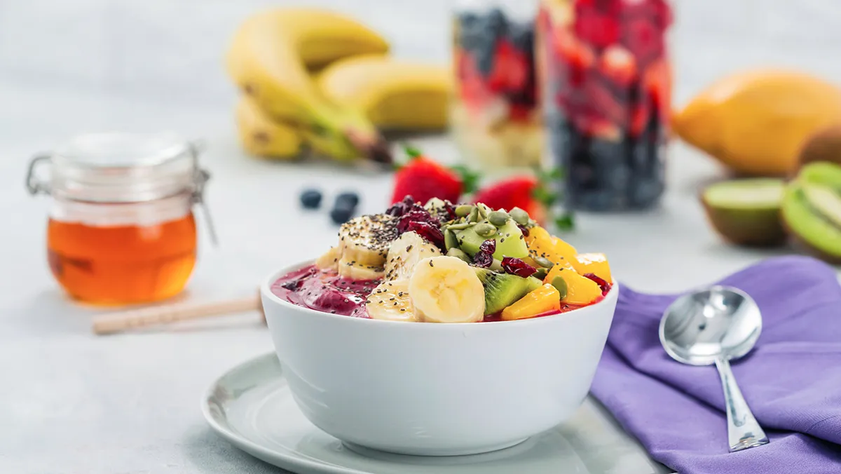 Un bol de fruits coloré avec des bananes, des fraises, des kiwis et de la mangue, arrosé de miel, sur une table avec d'autres fruits et une cuillère.