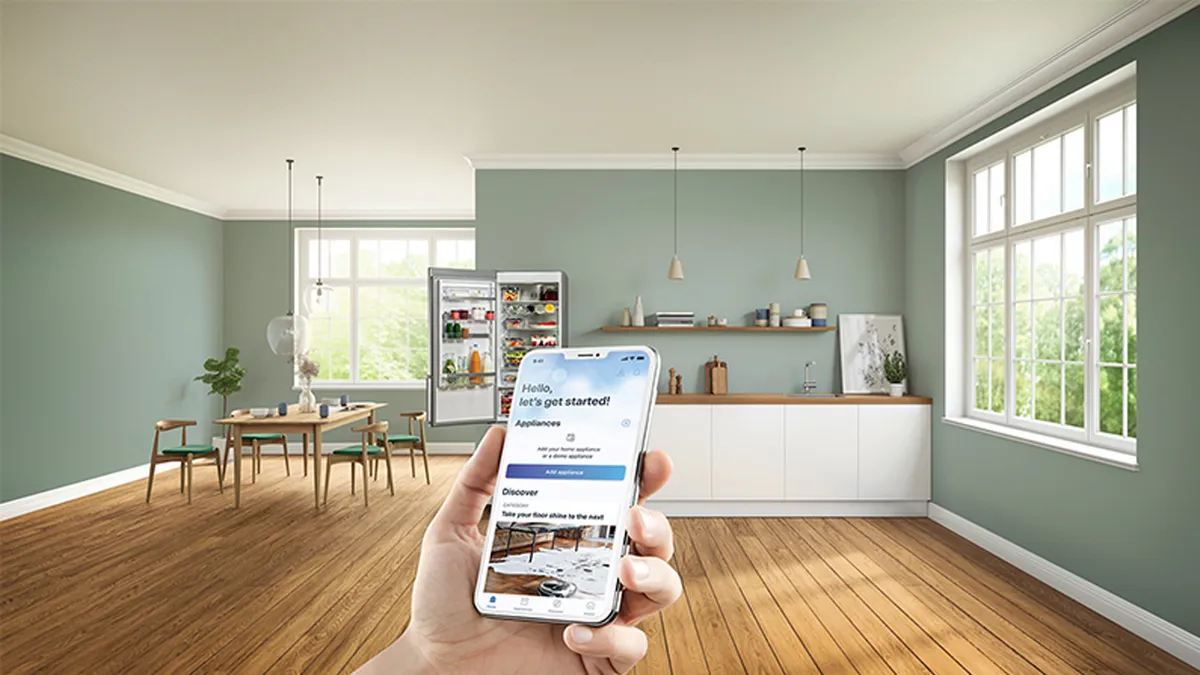 Eine Hand halt ein Smartphone mit einer angezeigten App in einer hellen, modernen KUche mit grünen WAnden, HolzbOden und einem Esstisch.
