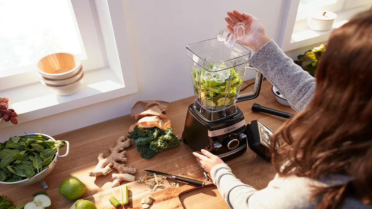 A person's hand interacts with the high performance blender's control panel and mixes the green ingredients.