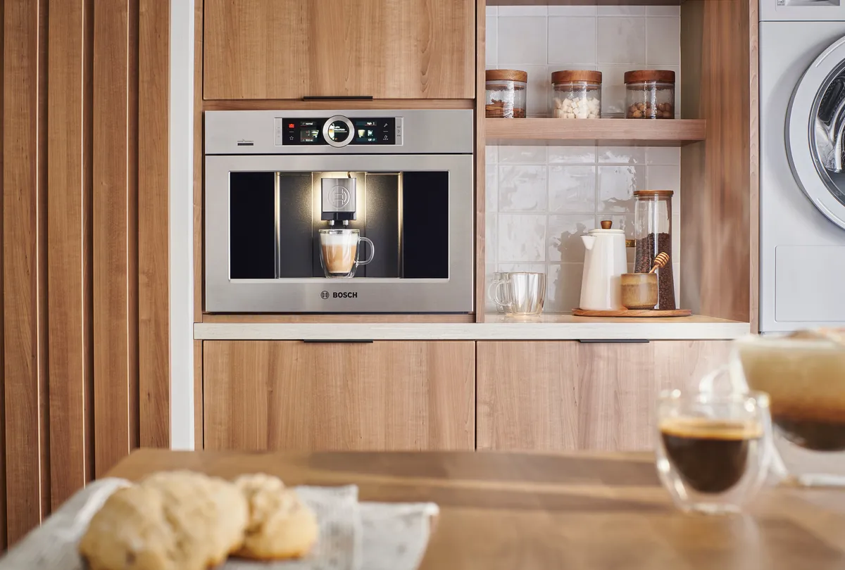 A modern Bosch coffee machine in a wooden kitchen, preparing coffee with cookies on a table in the foreground.