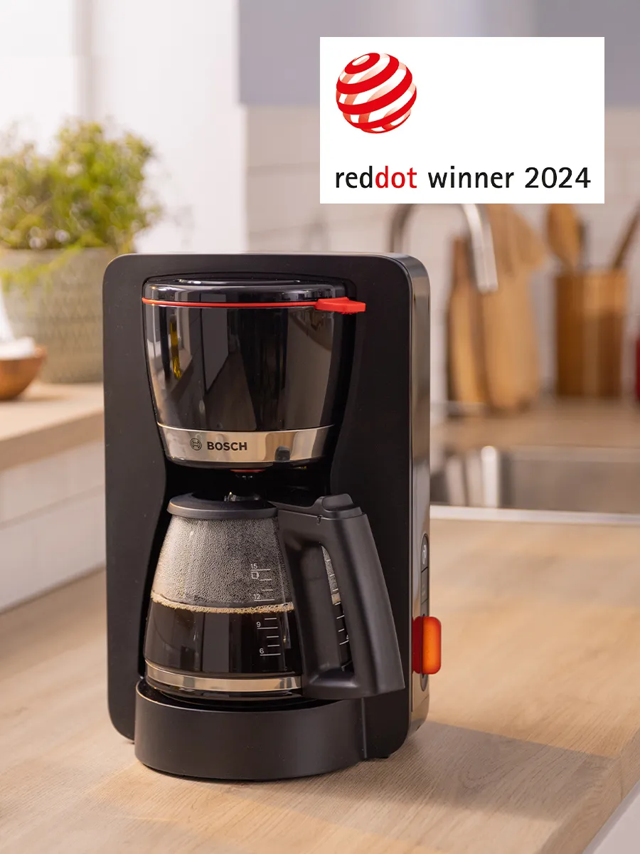 آلة صنع القهوة بالتنقيط من بوش MyMoment باللون الأسود مع إبريق زجاجي، حاصلة على جائزة Red Dot Winner 2024. تظهر الآلة على سطح مطبخ خشبي مع خلفية مطبخ غير واضحة.