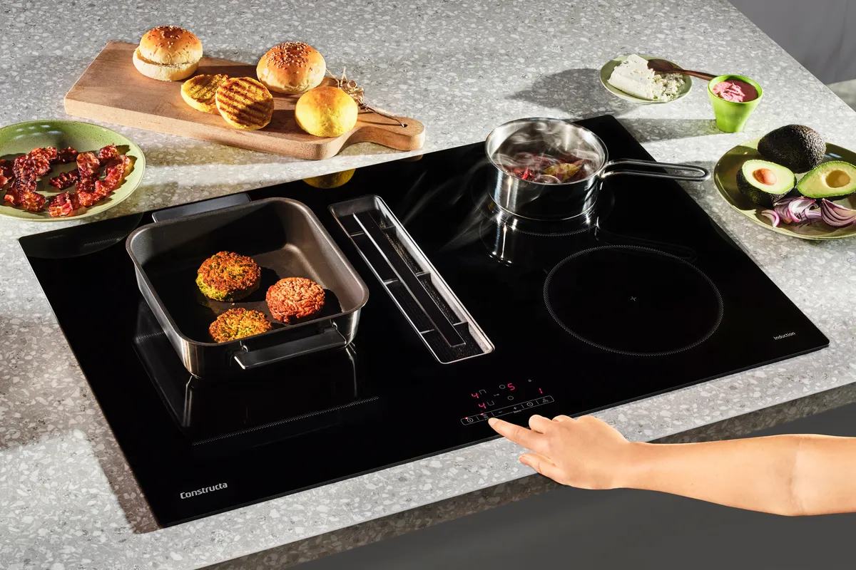 Ein modernes Induktionskochfeld mit einer Pfanne, die Gemüsepatties enthält, einem dampfenden Topf und frisch gebackenem Brot auf einem Holzbrett.