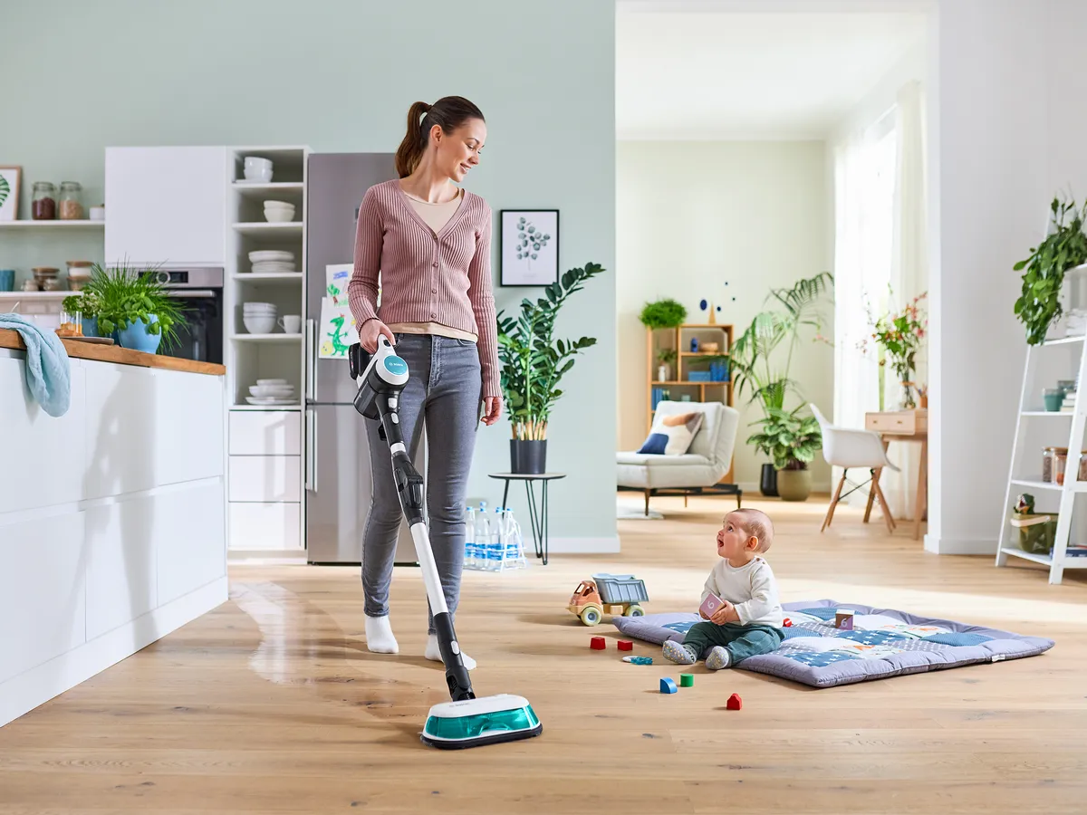 Kobieta odkurzająca salon, podczas gdy dziecko bawi się na podłodze, otoczona roślinami i domowym wystrojem.