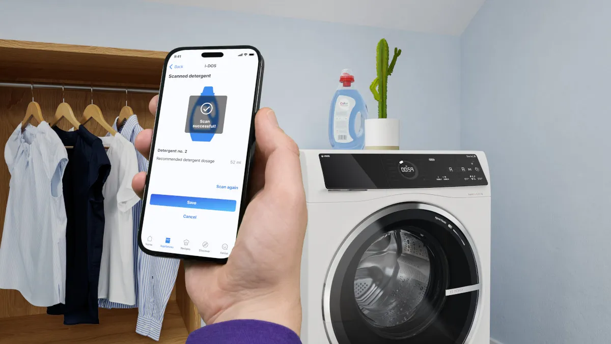 Eng Persoun hält en Smartphone, deen eng Wäsche-App weist, während se nieft enger Wäschemaschinn an engem Kleederschaf stécht.